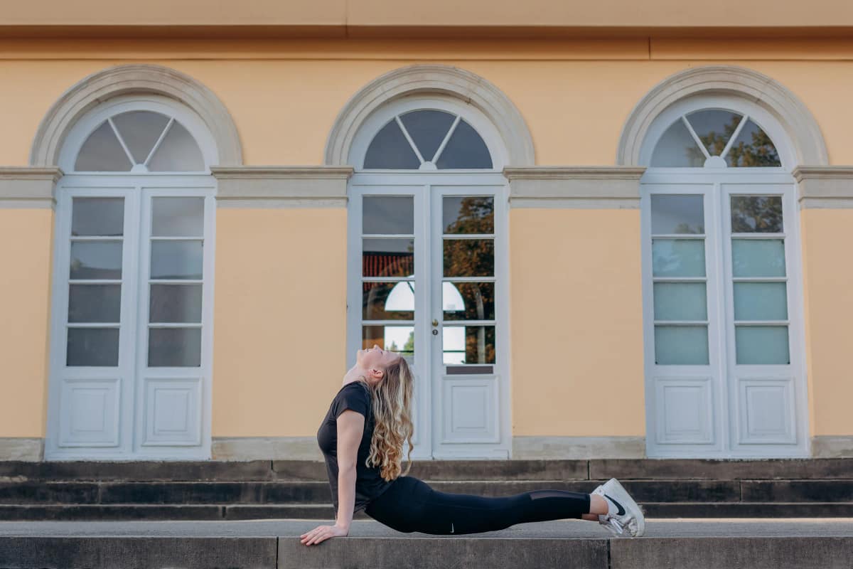 Personal Trainerin Lara Kölzow in der Yoga-Haltung Kobra auf einer Steinstufe vor einem gelben Gebäude mit weißen Bogenfenstern